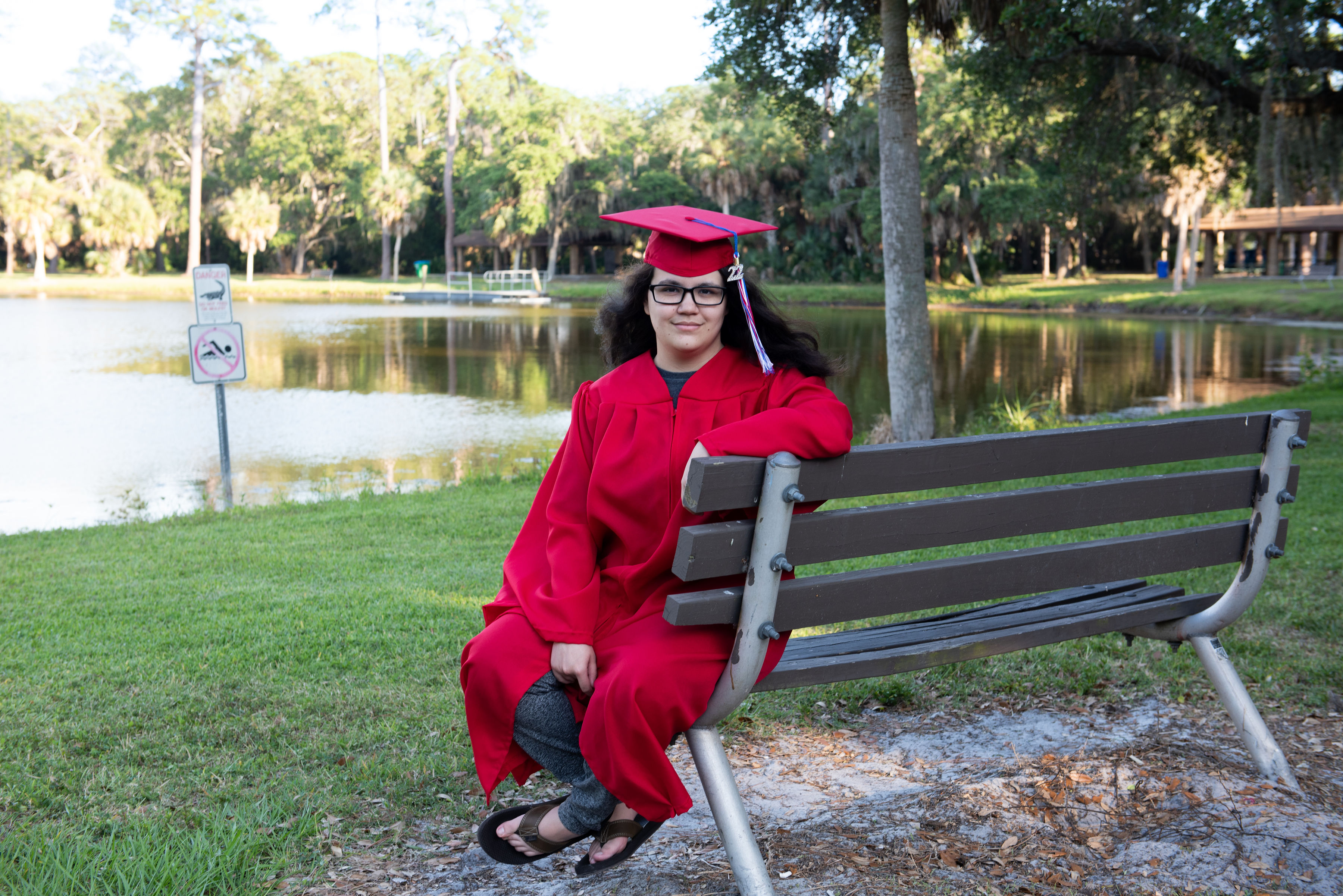 Senior sitting on park bench by lake with graduation cap in Florida woodland