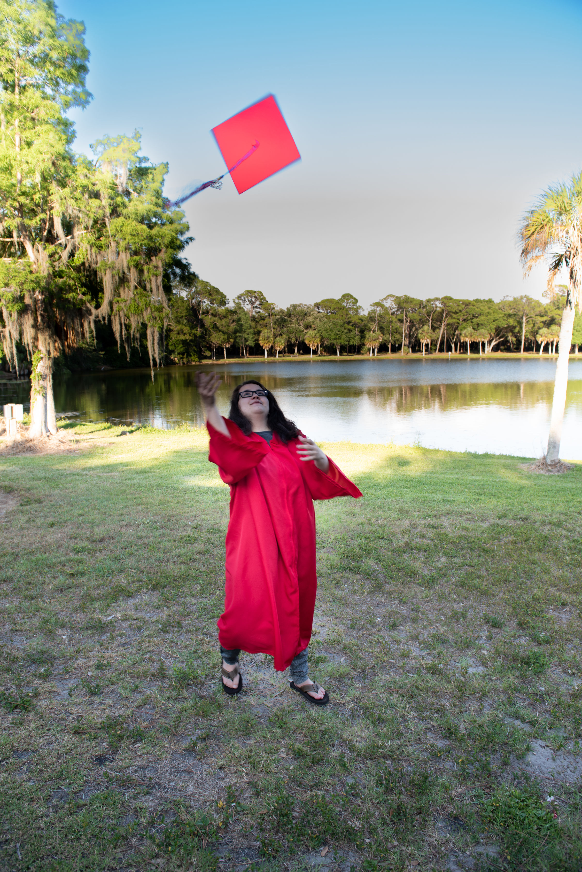 Senior tossing graduation cap by lake with Spanish moss trees in background